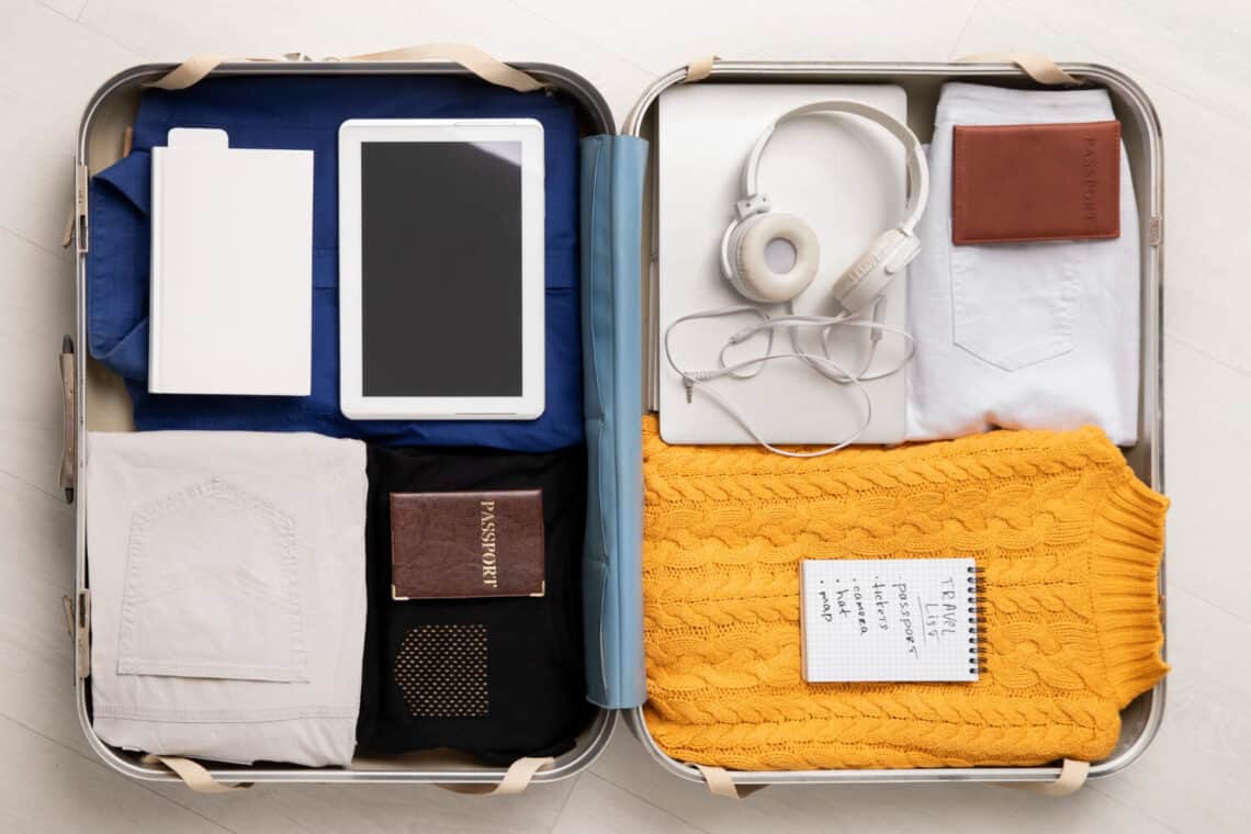 open luggage flat on the floor with ipad, head phones, clothes, yellow sweater, passport, list and travel essentials